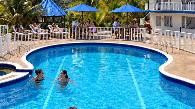 Personas nadando en la piscina de Hotel Cabaleón con parasoles azules y palmeras, pasadía en Tena